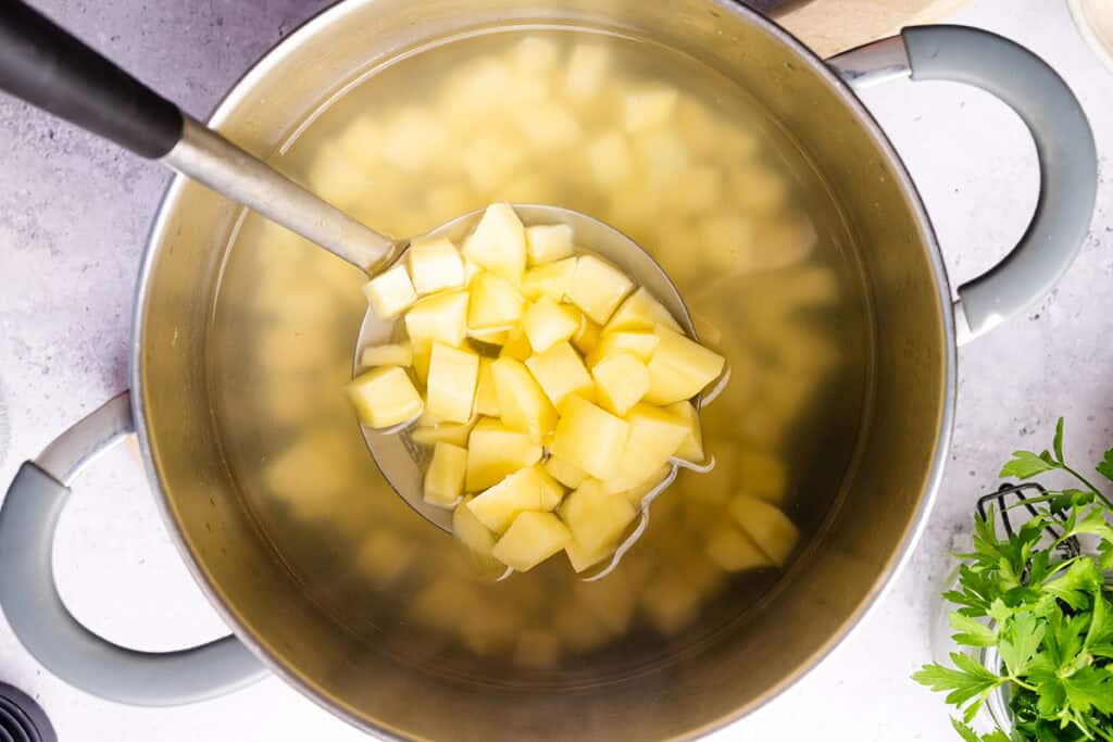 A ladle lifts diced potatoes from a large pot of water, seen from above—a classic step in prepping Homemade Potato Salad. Fresh parsley and a chopping board rest nearby on the white countertop.