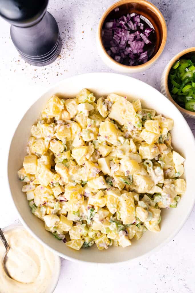 A bowl of creamy homemade potato salad with chopped green onions and red onions, surrounded by small bowls of sliced green onions, diced red onions, and a dish of mayonnaise on a light countertop.