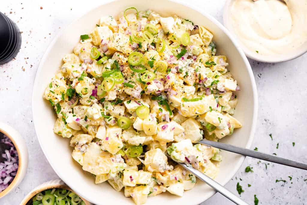 A bowl of creamy homemade potato salad garnished with chopped green onions, red onions, herbs, and black pepper, with two serving utensils in the bowl. Small bowls of ingredients are visible nearby.