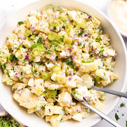 A bowl of creamy homemade potato salad garnished with chopped green onions, red onions, herbs, and black pepper, with two serving utensils in the bowl. Small bowls of ingredients are visible nearby.