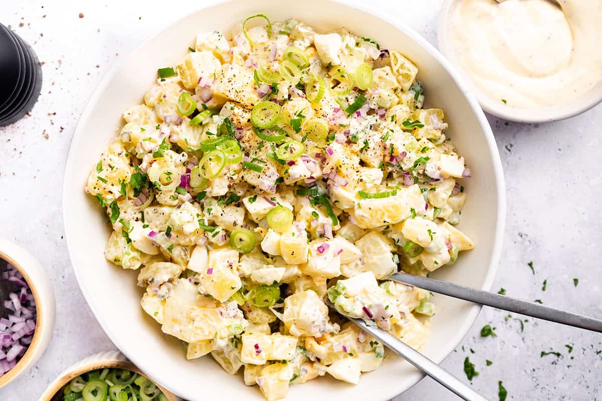 A bowl of creamy homemade potato salad garnished with chopped green onions, red onions, herbs, and black pepper, with two serving utensils in the bowl. Small bowls of ingredients are visible nearby.