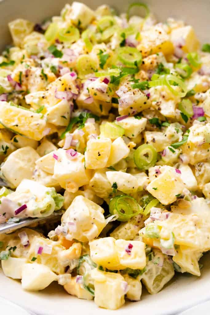 A close-up of a bowl of Homemade Potato Salad with diced potatoes, chopped green onions, red onions, celery, and herbs, all coated in a creamy dressing. A spoon is partially visible in the salad.