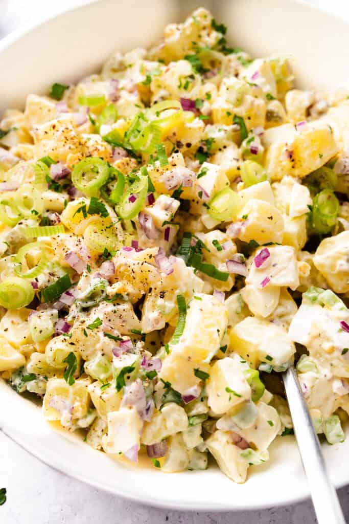 A close-up of a bowl of creamy Homemade Potato Salad garnished with chopped green onions, red onions, and fresh herbs, with a spoon resting in the mixture. The salad features visible chunks of potatoes and a pepper seasoning.