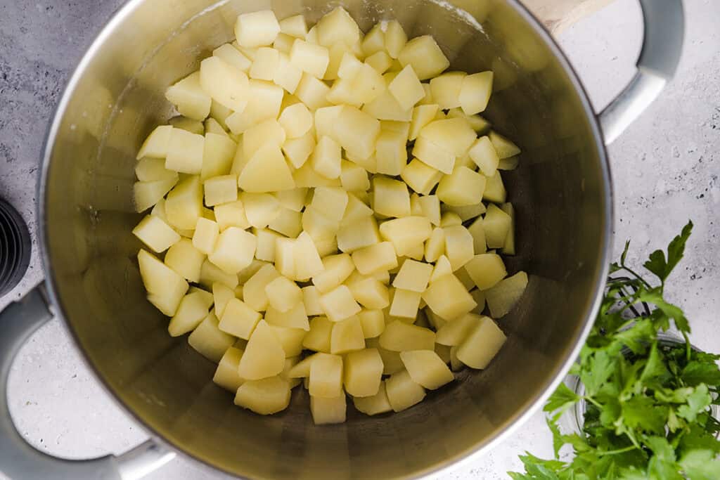 A large metal pot filled with peeled and diced raw potatoes sits on a light countertop, ready for homemade potato salad, with fresh green herbs visible nearby.
