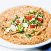 A bowl of refried beans topped with jalapenos, tomatoes, and crumbled cheese.