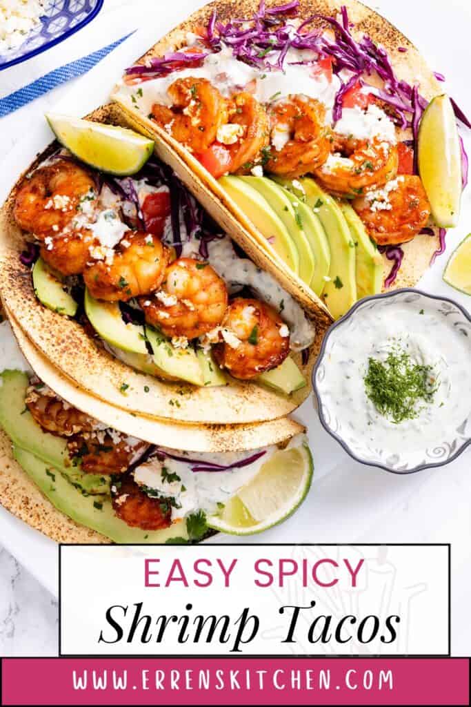 A close-up photo of two shrimp tacos on a white plate. The tacos are filled with seasoned shrimp, sliced avocado, shredded cabbage, and a white crema sauce. Text overlay on the plate reads "Easy Spicy Shrimp Tacos"