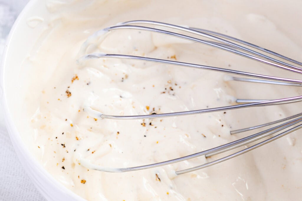 A metal whisk resting in a bowl of creamy, white sauce speckled with ground black pepper—just what you need for the Best Classic Egg Salad Recipe.