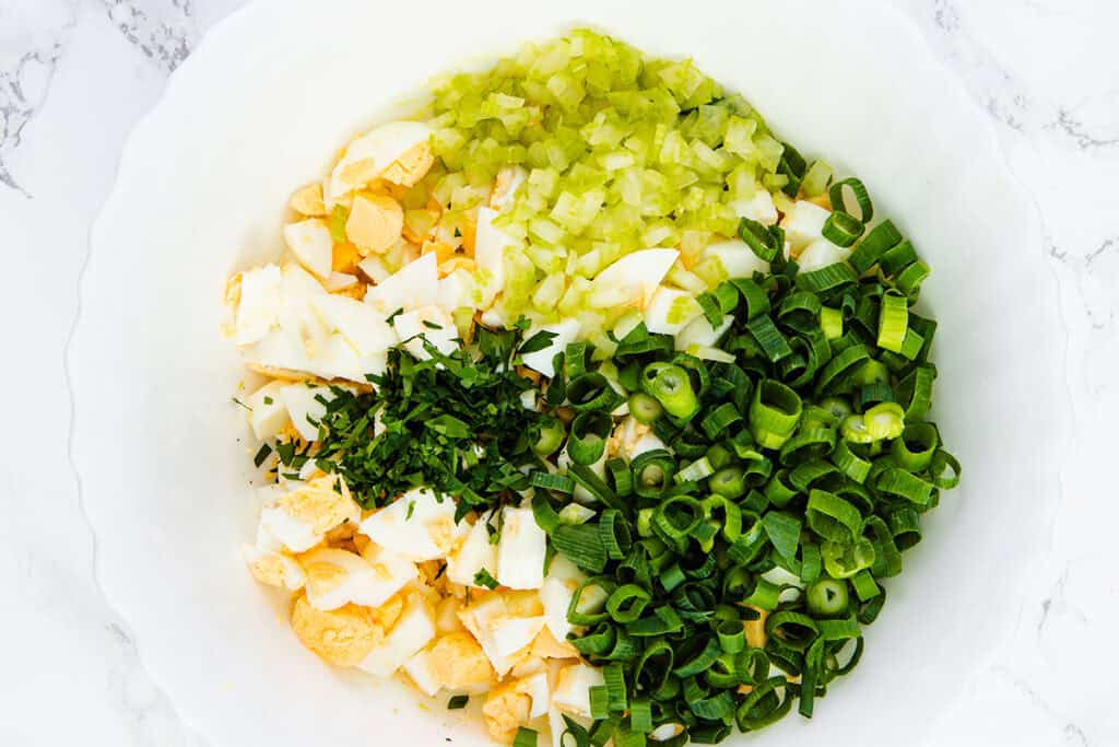 A white bowl containing chopped hard-boiled eggs, celery, scallions, and parsley arranged in sections, ready to be mixed for the Best Classic Egg Salad Recipe.