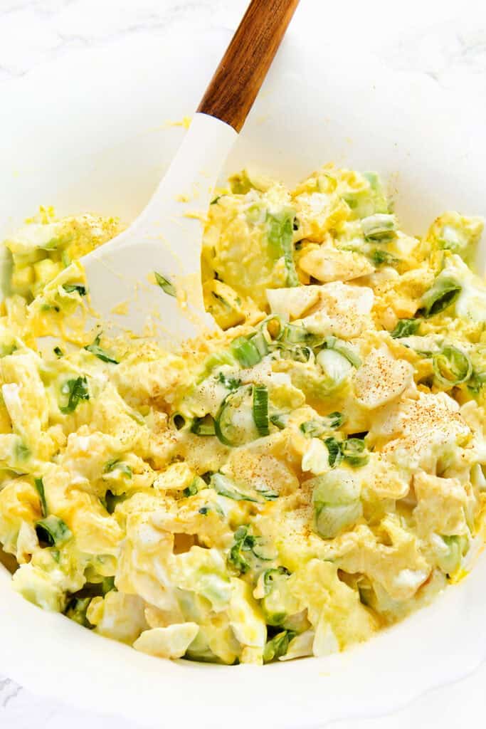 A white bowl of the Best Classic Egg Salad Recipe with creamy egg salad, chopped green onions, sprinkled with paprika, and a white spatula with a wooden handle resting inside.