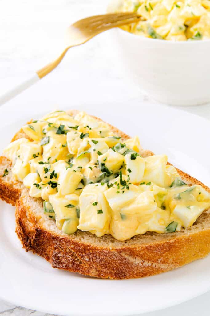 A slice of rustic bread topped with creamy egg salad, garnished with chopped herbs—an inviting plate that showcases the Best Classic Egg Salad Recipe, served with extra salad and a fork in the background.