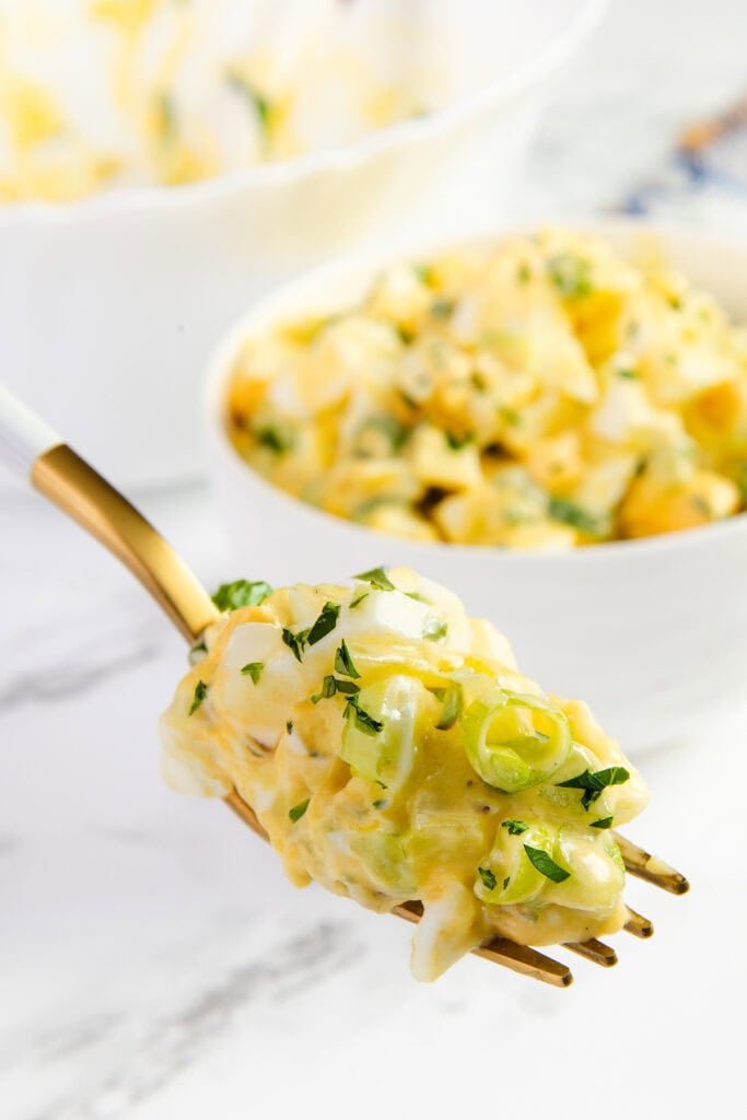 A close-up of a forkful of creamy potato salad with chopped green onions and parsley, reminiscent of the Best Classic Egg Salad Recipe, held in front of a white bowl filled with more potato salad on a bright, white surface.