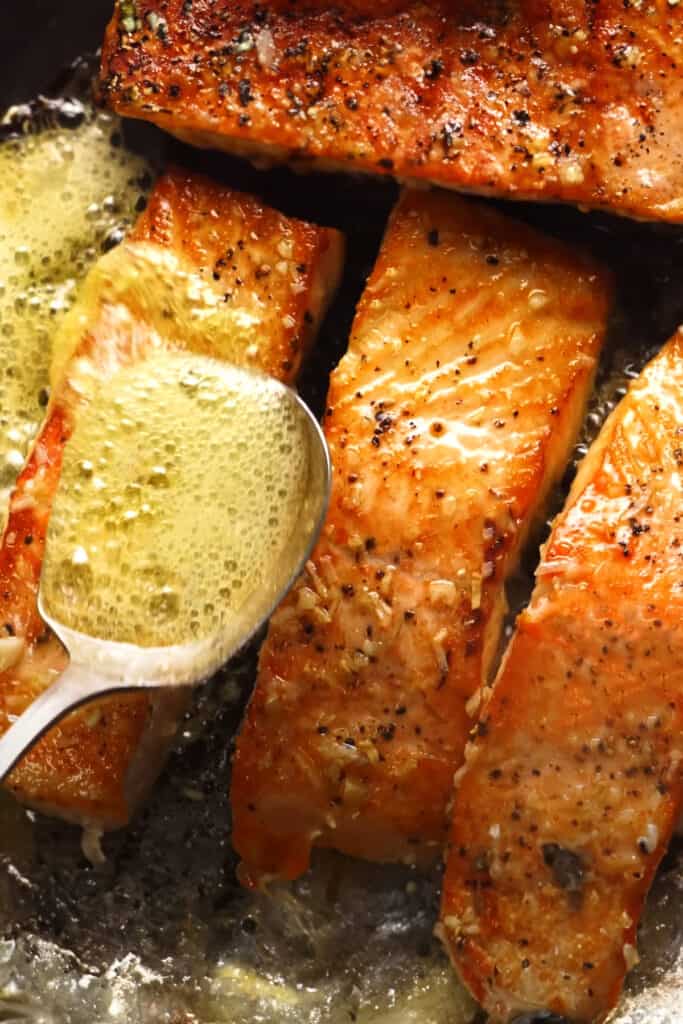 a spoon pouring butter onto salmon cooking in a pan.