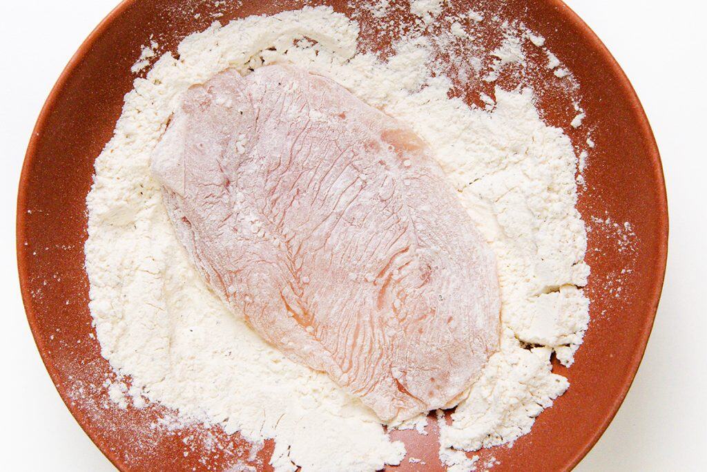 A raw piece of meat, perfect for Chicken Marsala, lies on a bed of white flour in a brown bowl, ready to be coated, against a white background.