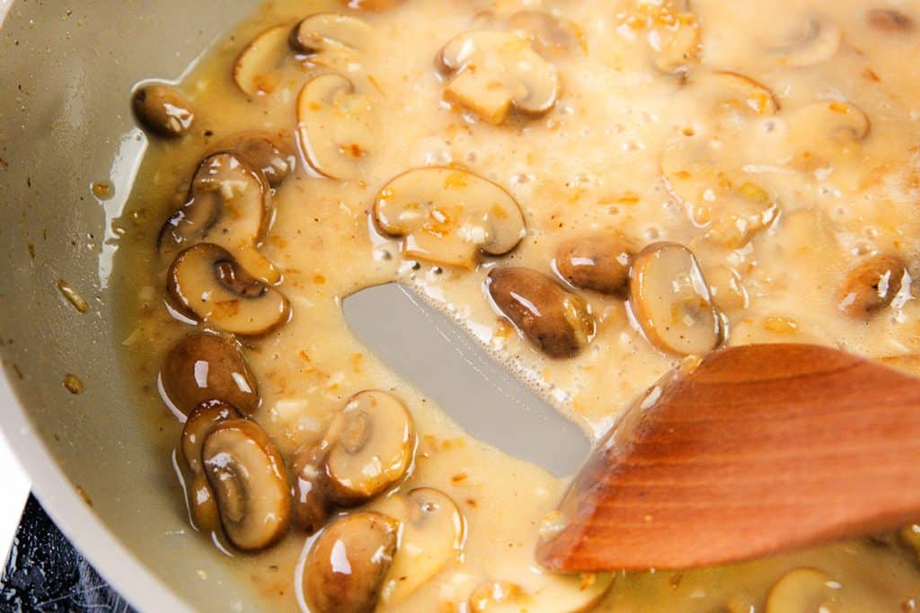 Sliced mushrooms cooking in a creamy Chicken Marsala sauce in a pan, being stirred with a wooden spatula.