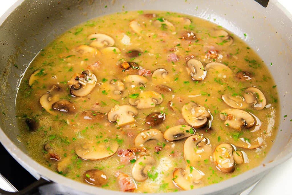 A close-up of a pan filled with simmering Chicken Marsala sauce, showing sliced mushrooms, chopped herbs, and small pieces of bacon in a light, brothy liquid.