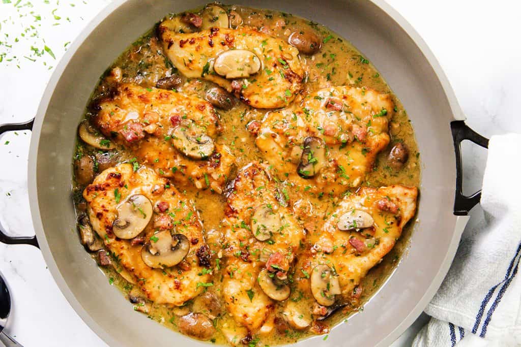A skillet filled with golden-brown Chicken Marsala breasts in a creamy sauce with mushrooms, bacon bits, and fresh herbs, viewed from above.