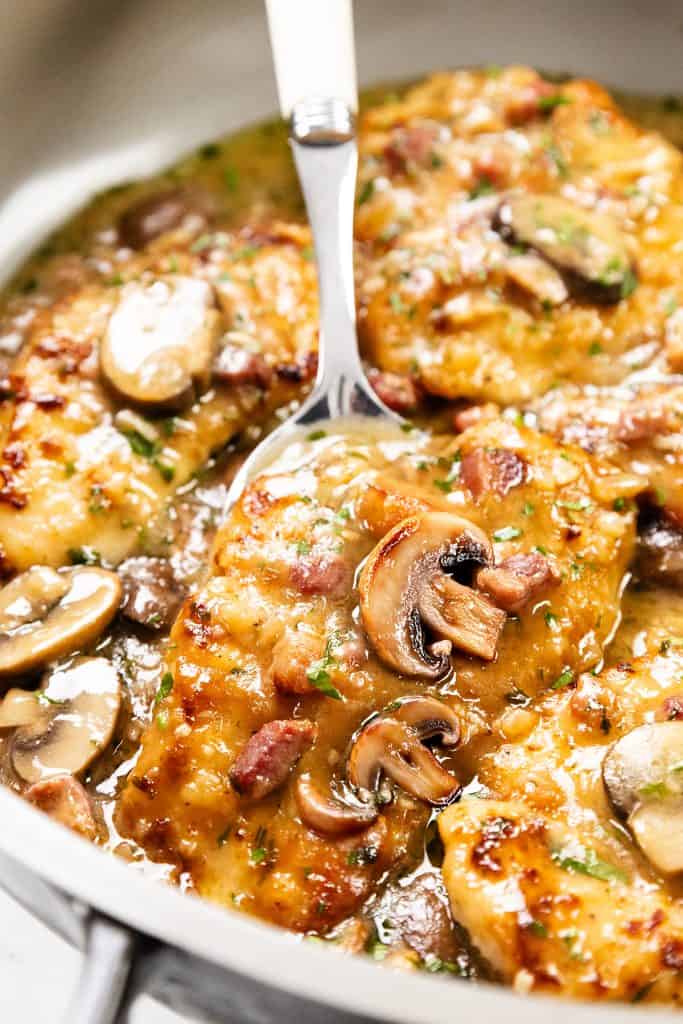 A close-up of Chicken Marsala in a pan, featuring golden-brown chicken breasts, sautéed mushrooms, and a rich marsala sauce. A serving spoon lifts a piece of the hearty, flavorful Chicken Marsala for serving.