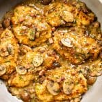 A skillet filled with golden-brown Chicken Marsala cutlets in a creamy sauce, topped with sliced mushrooms and herbs, with bits of bacon and whole mushrooms visible in the pan.