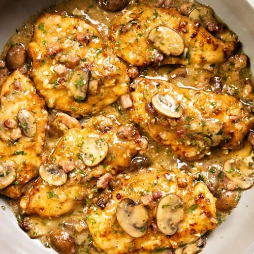 A skillet filled with golden-brown Chicken Marsala cutlets in a creamy sauce, topped with sliced mushrooms and herbs, with bits of bacon and whole mushrooms visible in the pan.