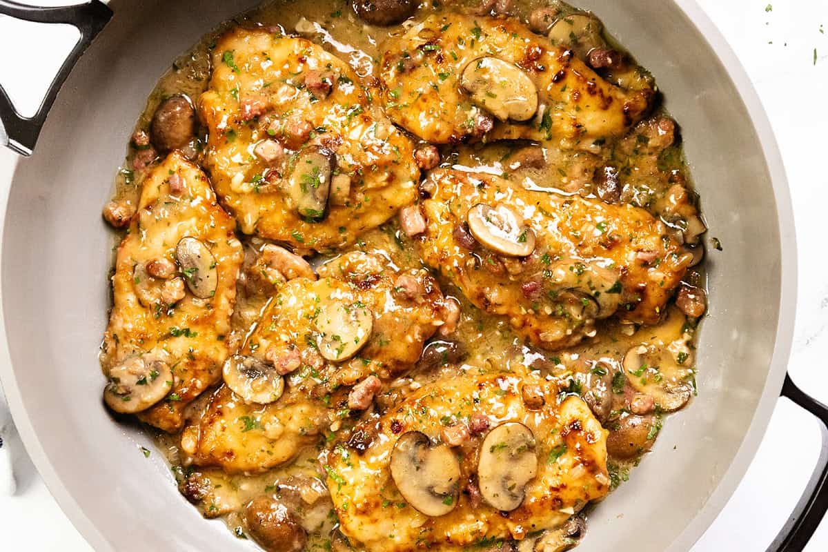 A skillet filled with golden-brown Chicken Marsala cutlets in a creamy sauce, topped with sliced mushrooms and herbs, with bits of bacon and whole mushrooms visible in the pan.
