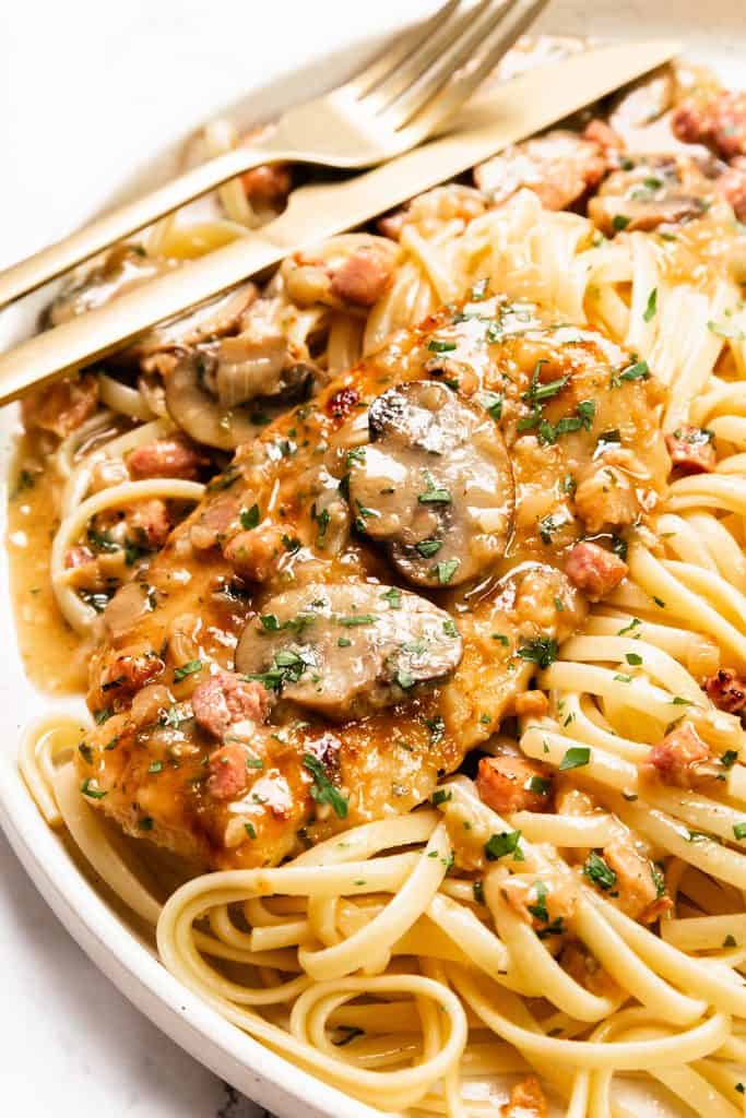 A plate of pasta topped with a breaded Chicken Marsala cutlet, creamy mushroom sauce, diced bacon, and chopped parsley, served with a gold fork and knife.