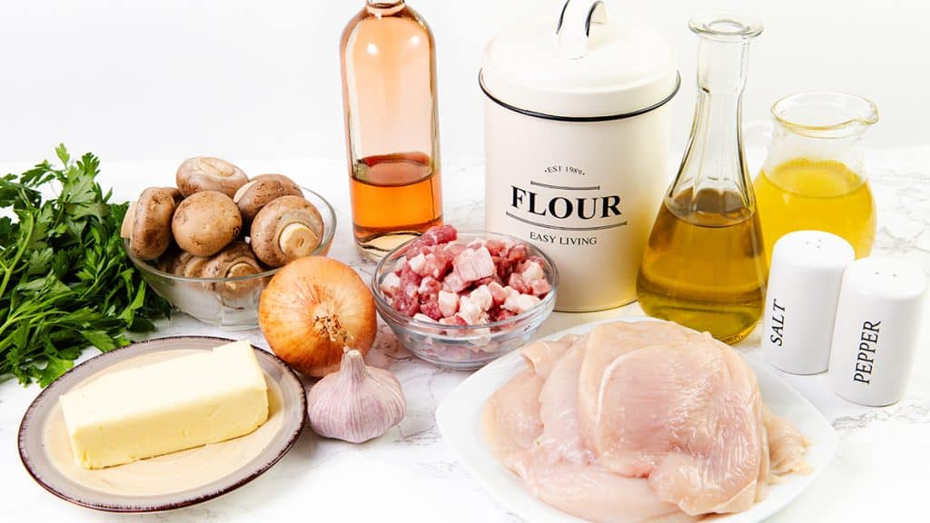 A variety of cooking ingredients on a white surface, including raw chicken breasts for Chicken Marsala, diced bacon, mushrooms, onion, garlic, parsley, butter, flour, oil, vinegar, and salt and pepper shakers.