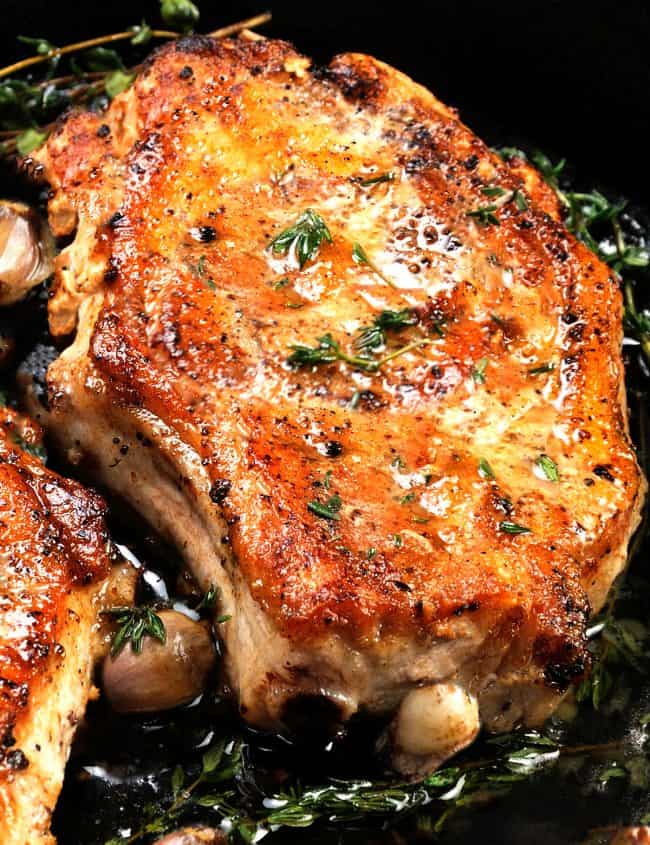 Juicy, browned pork steak on a bone in oil with garlic and herbs in a frying pan. Close-up.
