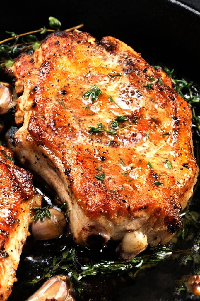 Juicy, browned pork steak on a bone in oil with garlic and herbs in a frying pan. Close-up.