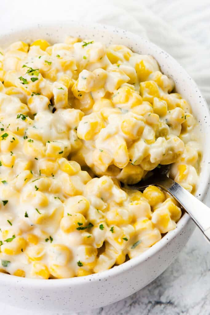 A bowl of creamed corn with a rich, white sauce infused with cream cheese. The dish is garnished with finely chopped herbs, appearing soft and luscious. A silver spoon rests inside the bowl, surrounded by a white textured background.