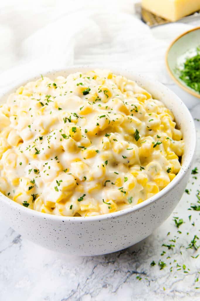 A bowl filled with creamy macaroni and cheese, accented by a touch of cream cheese corn and topped with chopped parsley. The speckled bowl rests on a marble surface, while a small dish with more parsley lingers in the background.