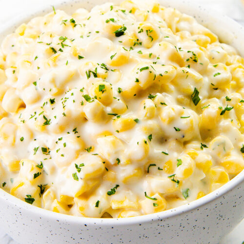 A bowl of creamy, cheesy creamed corn topped with chopped herbs. The dish appears rich and indulgent, with a smooth sauce coating the corn kernels. The bowl is white with small speckles, sitting on a light marble surface.