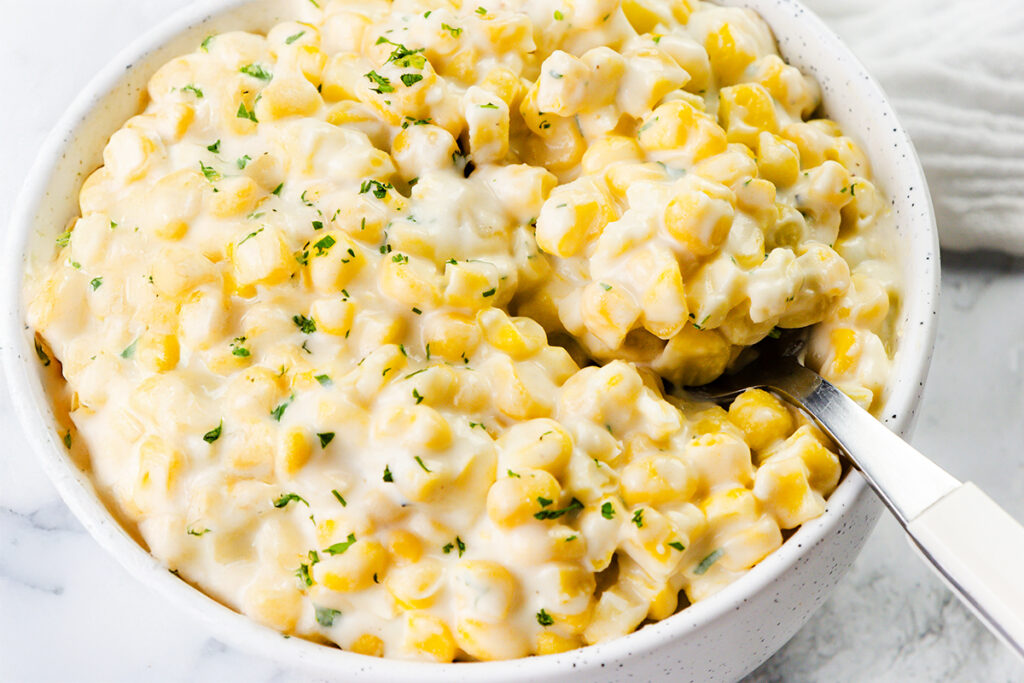 A bowl of creamy corn salad, enriched with cream cheese and topped with finely chopped parsley, sits invitingly on a white marble surface. The rich, thick texture showcases succulent corn kernels immersed in a luscious sauce. A spoon rests in the bowl, ready for serving.