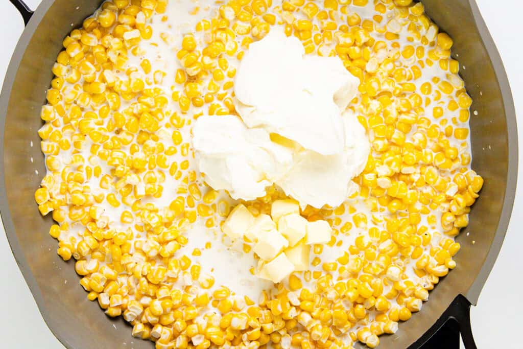 A pan filled with corn kernels, milk, cubes of butter, and a touch of cream cheese awaits a swirl that promises a delightful Cream Cheese Corn dish. The bright, fresh ingredients contrast beautifully against the dark rim of the pan.