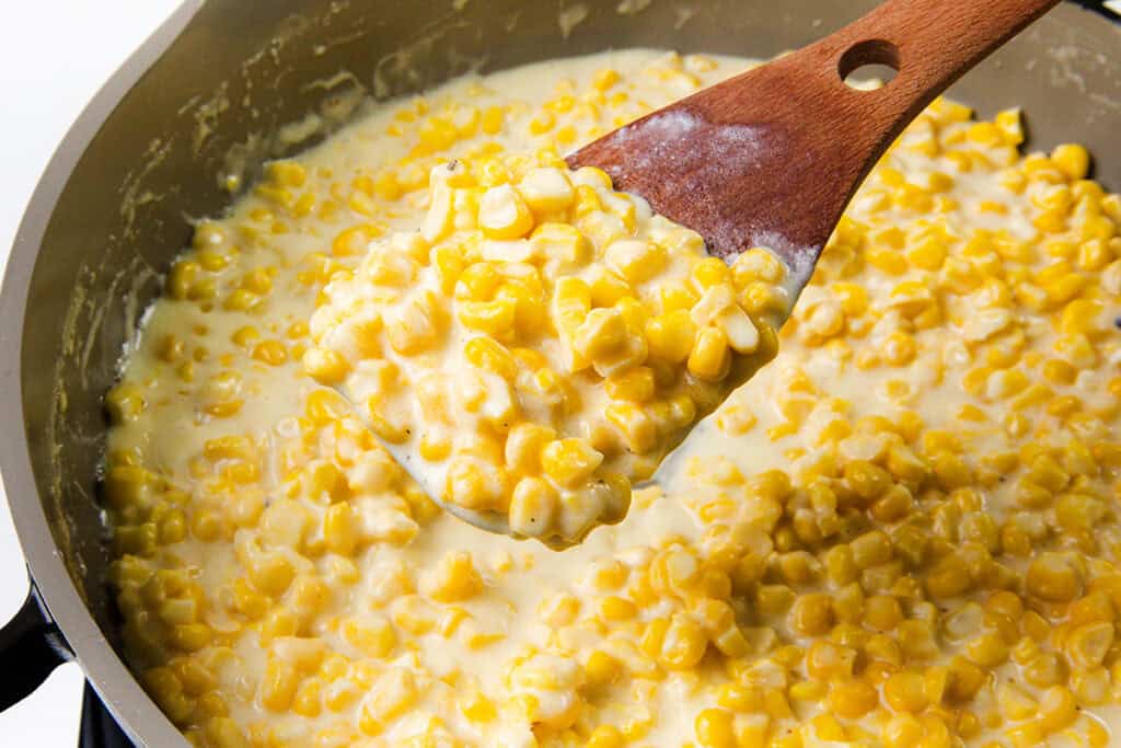 A wooden spoon scoops up creamy corn from the pot, revealing vibrant yellow kernels swirled into a luscious cream cheese sauce. This Creamed Corn dish boasts a rich, thick appearance that promises indulgence in every bite.