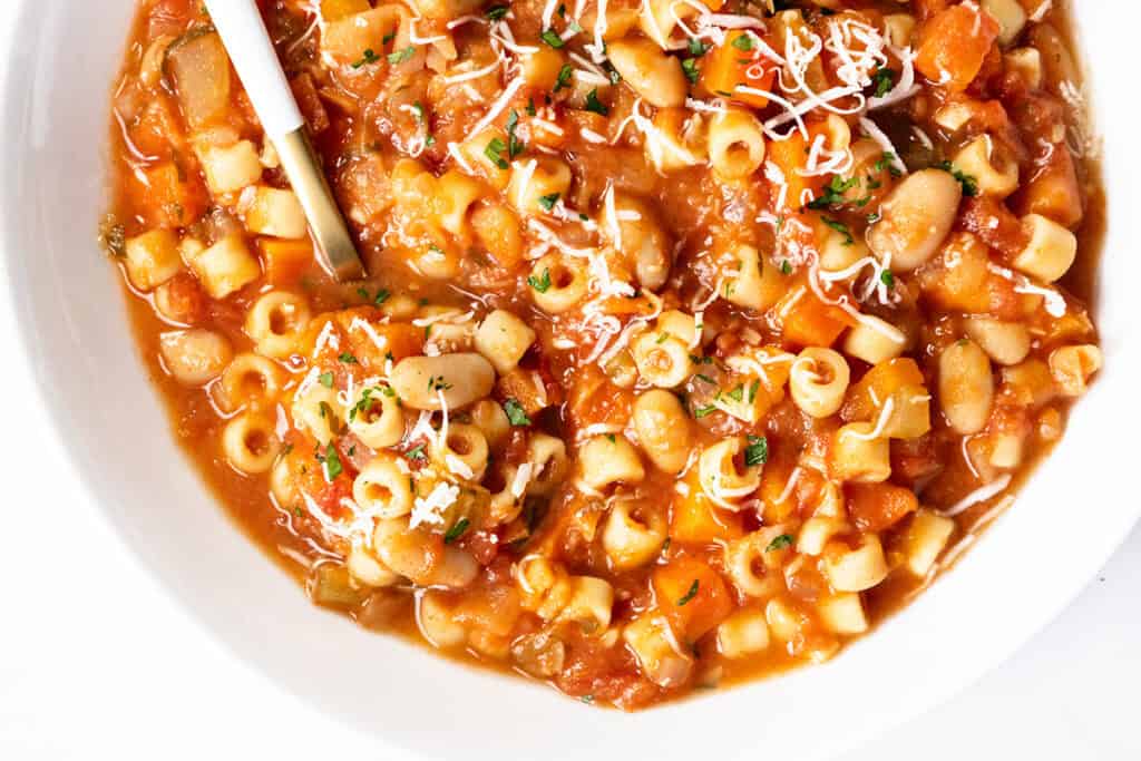 A close-up of Pasta E Fagioli Soup featuring ditalini pasta, white beans, tomatoes, and vegetables, all garnished with grated cheese and chopped herbs, with a spoon resting in the bowl.