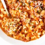 A close-up of Pasta E Fagioli Soup featuring ditalini pasta, white beans, tomatoes, and vegetables, all garnished with grated cheese and chopped herbs, with a spoon resting in the bowl.