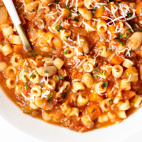 A close-up of Pasta E Fagioli Soup featuring ditalini pasta, white beans, tomatoes, and vegetables, all garnished with grated cheese and chopped herbs, with a spoon resting in the bowl.