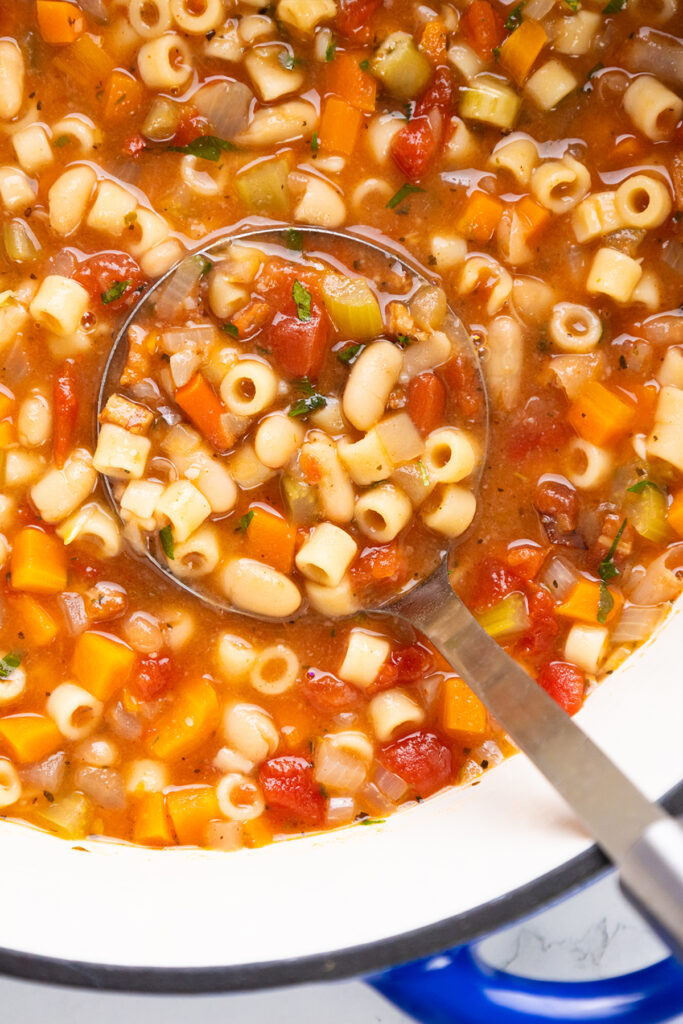 A close-up of a pot of Minestrone or Pasta E Fagioli Soup, with a ladle lifting a serving. The soup features ditalini pasta, white beans, diced carrots, celery, tomatoes, and herbs in a savory tomato-based broth.