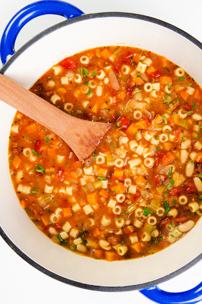 A large white pot with blue handles holds hearty Pasta E Fagioli Soup, brimming with ditalini pasta, vegetables, beans, and herbs. A wooden spoon rests in the vibrant, flavorful mix.