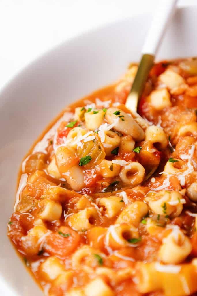 Close-up of a bowl of Pasta E Fagioli Soup with ditalini pasta, tomatoes, beans, and vegetables, garnished with grated cheese and chopped herbs, with a spoon dipped into the hearty soup.
