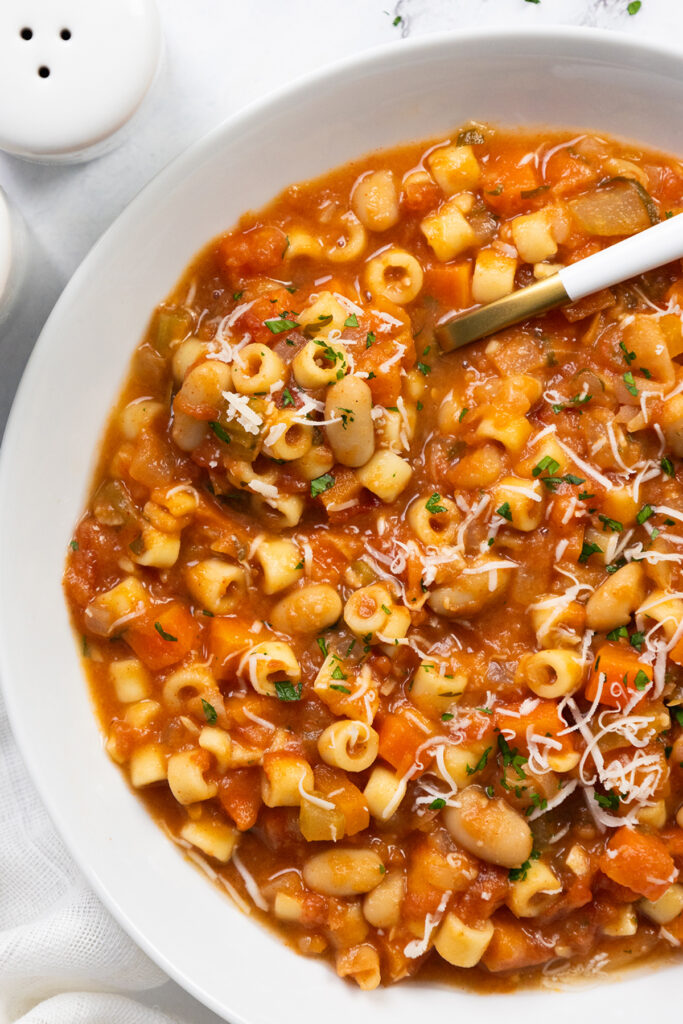 A close-up of Pasta E Fagioli Soup shows small pasta, white beans, and vegetables in a rich tomato broth, garnished with grated cheese and chopped herbs, with a spoon on the side.