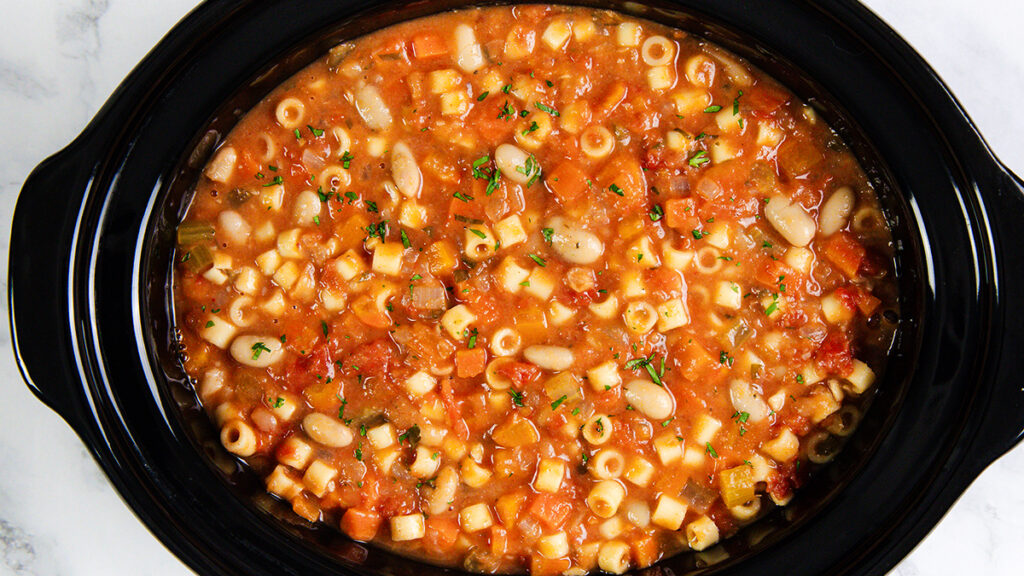 A slow cooker filled with hearty Pasta E Fagioli Soup featuring ditalini pasta, beans, diced tomatoes, carrots, and herbs in a rich tomato-based broth.