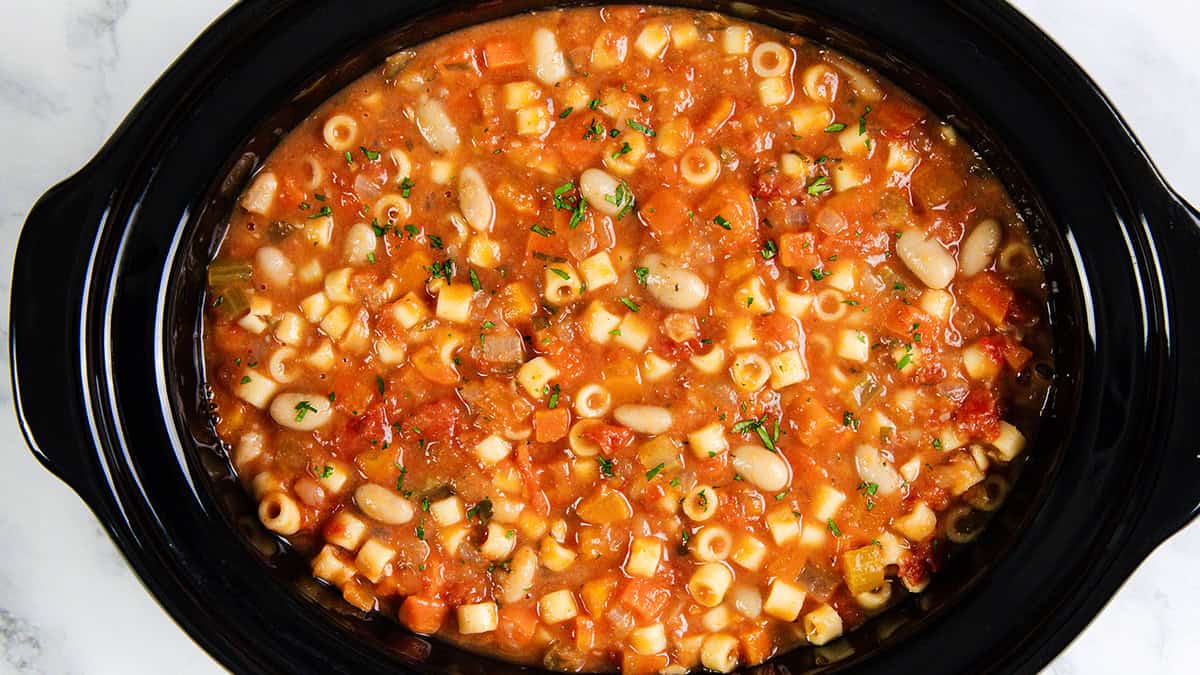 A slow cooker filled with hearty Pasta E Fagioli Soup featuring ditalini pasta, beans, diced tomatoes, carrots, and herbs in a rich tomato-based broth.