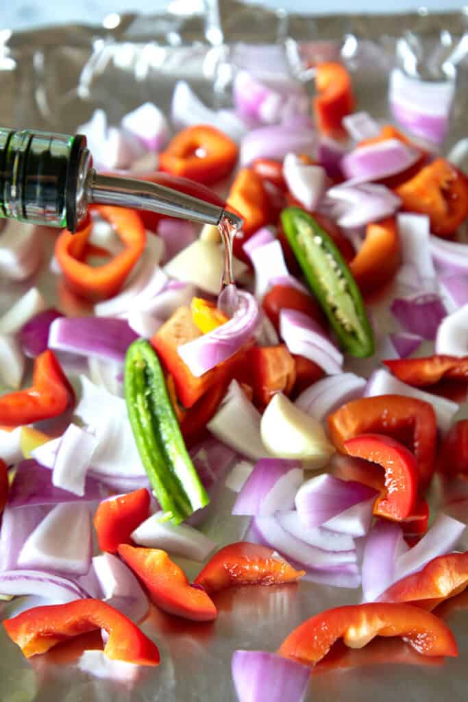 the onions, garlic, and peppers on a sheet pan being drizzled with oil.