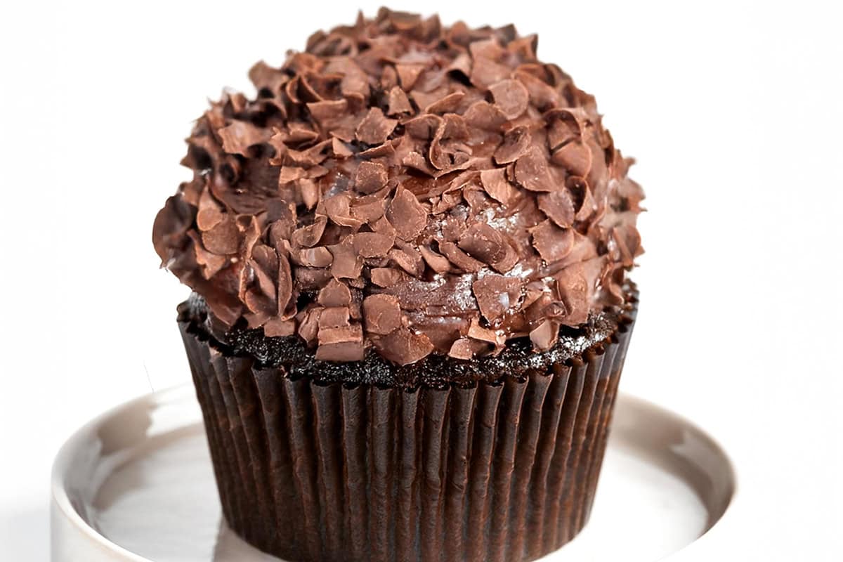 A luscious chocolate cupcake with creamy frosting and delicate chocolate shavings graces a white surface. Nestled in a dark brown paper liner, this delectable treat epitomizes the rich indulgence of classic Chocolate Cupcakes.