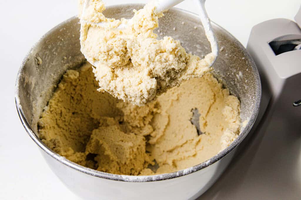 A stand mixer bowl filled with thick cookie dough, with some dough clinging to the attached paddle above the bowl. The mixer is partially visible on the side against a white background, perfect for whipping up the Best Lemon Squares Recipe.