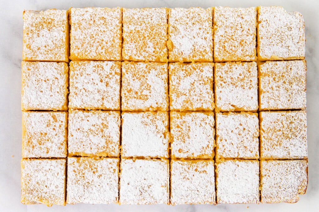 Twenty-four neatly cut lemon bars, made from the Best Lemon Squares Recipe and dusted with powdered sugar, are arranged in a rectangular grid on a white surface.