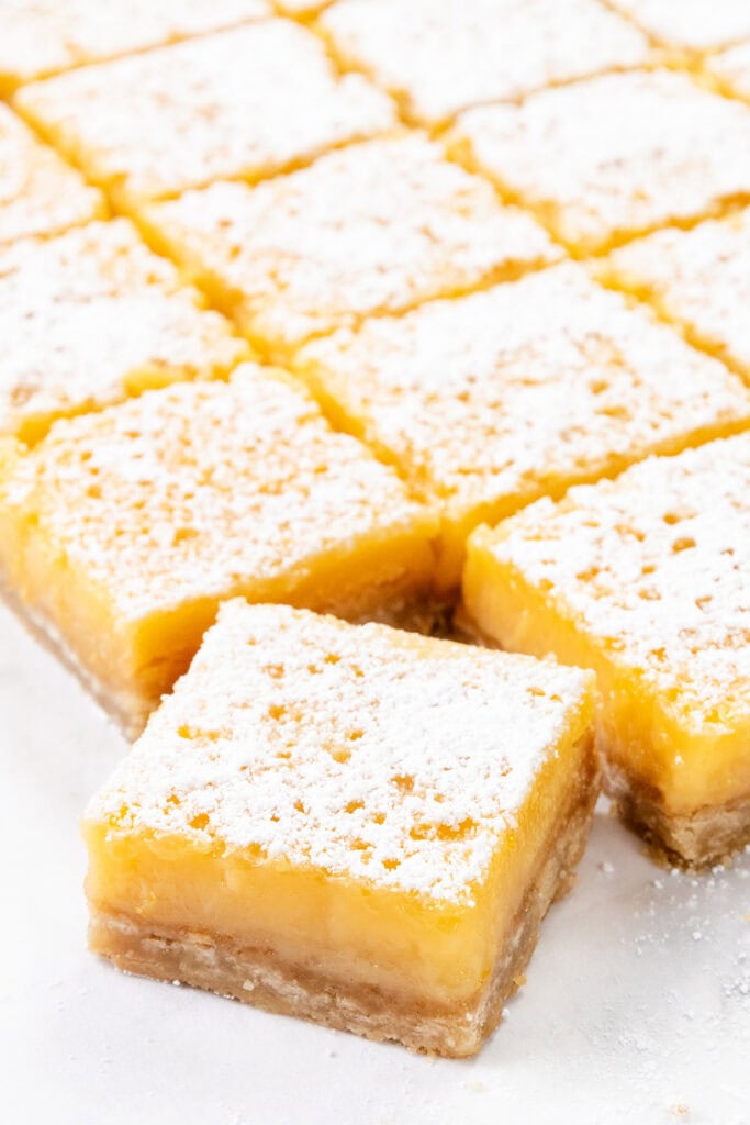 Close-up of several lemon bars from the Best Lemon Squares Recipe, topped with powdered sugar, cut into neat squares and arranged on a white surface, with one bar slightly pulled forward to show its layers.