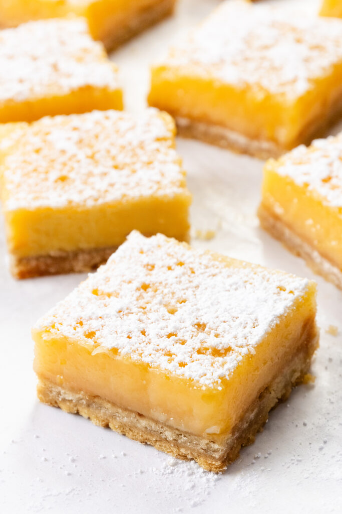 Several lemon bars, made from the Best Lemon Squares Recipe, are arranged on a white surface. Each bar features a thick yellow lemon filling and crumbly crust, topped with a generous dusting of powdered sugar.