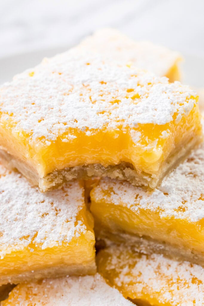 A close-up of stacked Lemon Bars with a dusting of powdered sugar on top. The bars have a golden-yellow, creamy filling and a bite taken out of one, revealing their soft, gooey texture.